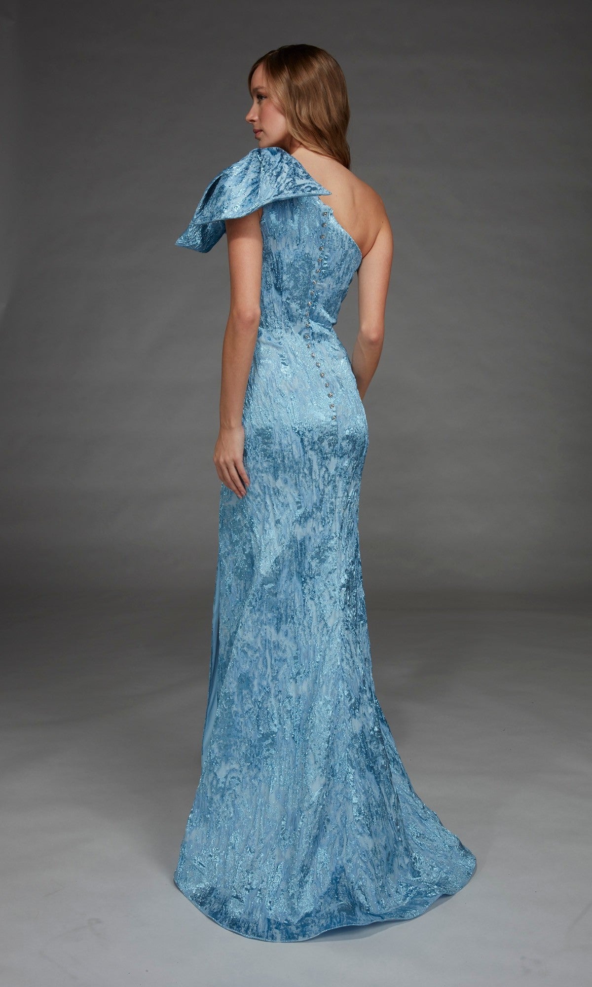 A woman is seen from the back in a studio, wearing the Alyce 27906 long formal dressโa light blue, floor-length gown with a textured fabric and a one-shoulder neckline.